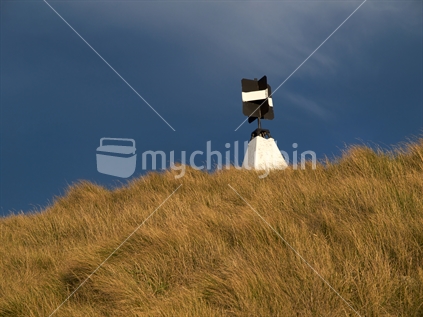 A New Zealand trig station in a long grass paddock, catching the evening sun against a stormy cloud background.