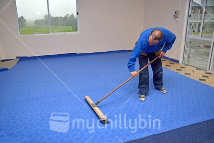 A floorlayer carefully uses a wide broom to smooth out the wrinkles in his newly laid carpet, over underlay.