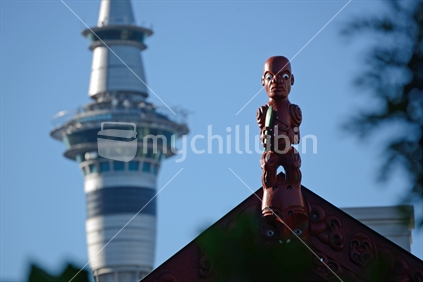 A carving of a Maori warrior stands guard over a Waipapa Marae while the modern Sky Tower dominates the Auckland skyline in New Zealand
