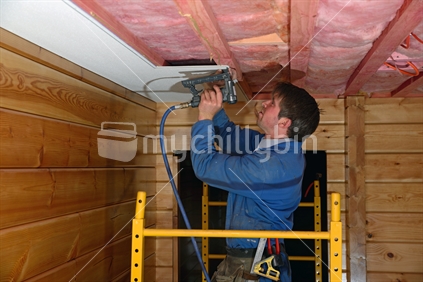 A builder staples up tiles in the ceiling of a lockwood style building