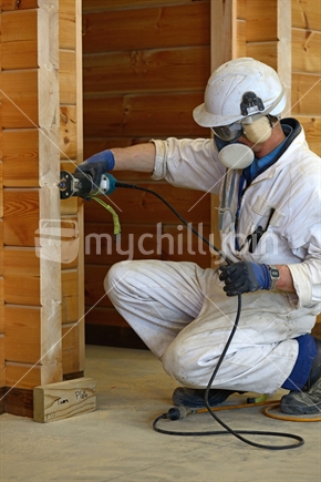 A painter in full protective clothing uses a hand-held router to round off corners on the wall of a lockwood style building before applying a clear top coat.