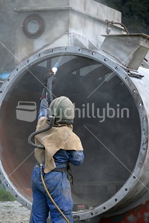 A man wearing full safety gear uses ground glass to sandblast the steel casing of a meat drying machine for a rendering plant