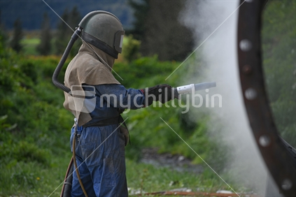 A man wearing full safety gear uses ground glass to sandblast the steel casing of a meat drying machine for a rendering plant