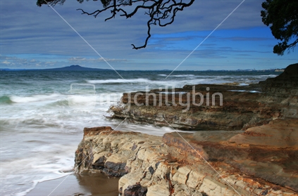 A rocky New Zealand coast swept by the wind, with iconic Rangitoto Island in the distance.  