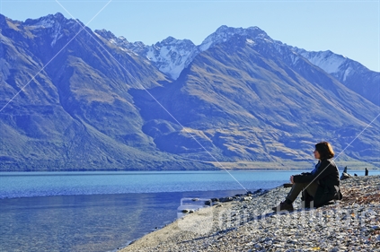 A woman sits on the stony shore of Lake Wakatipu looking across to the snow-capped mountains, Queenstown.