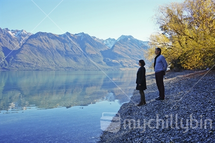 A couple stand together on the stony shores of Lake Wakatipu in autumn, enjoying the view