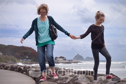A mother and daughter walking on foreshore rocks whilst holding hands.