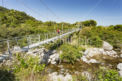 Red backpacker traverses swing bridge over river. Heaphy Track great walk, Kahurangi National Park
