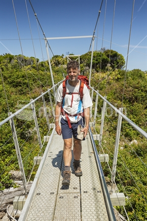 Kiwi man tramps over swing bridge over river on historic Heaphy Track great walk, Kahurangi National Park