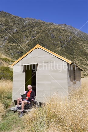 Kiwi male tramper sits in doorway of DOC hut at Top Waitaha, West Coast mountain wilderness