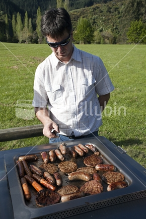 NZ male (in sunglasses) frying sausages and meat patties on a barbeque.