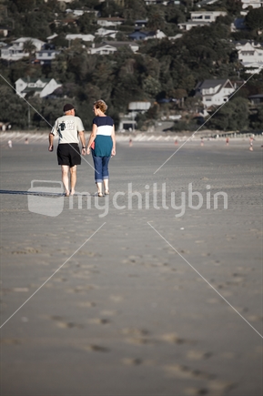 Elderly couple walk down sandy beach, Nelson.  Old guys rule tea shirt. 