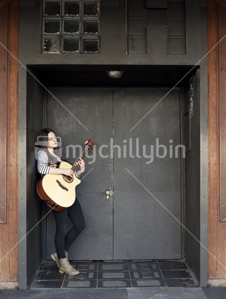Teenage girl playing guitar, in dark entrance of urban night club, with broken glass overhead.