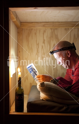 Male tramper reads by candlelight and headlight in a backcountry hut. Old Ghost Road, Buller region of West Coast.