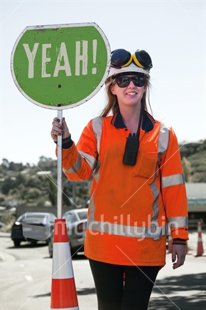 Young female traffic controller in hardhat and earmuffs holds YEAH! sign
