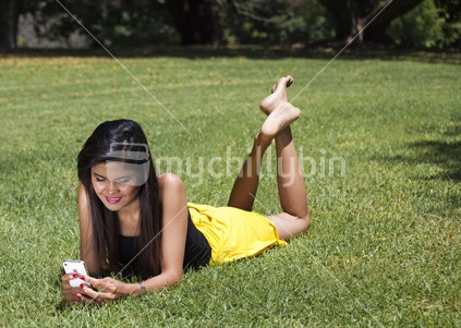 Young asian woman checks her cellular phone, while lying on green grass