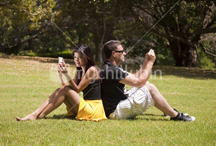 The Digital Divide. Ethnically diverse married couple sit back to back on grass while using their mobile phones