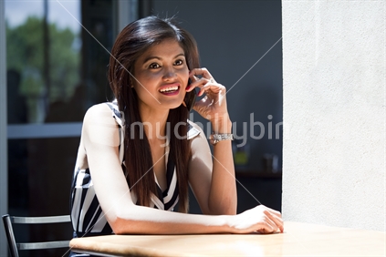 Attractive Asian lady seated at a cafeteria table enjoys talking on her phone