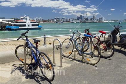 Bicycles and a scooter parked up by Devonport ferry terminal with a ferry and Auckland City CBD in the background.