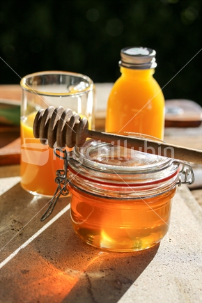 Jar of honey in the sunshine with glass and bottle of orange juice.