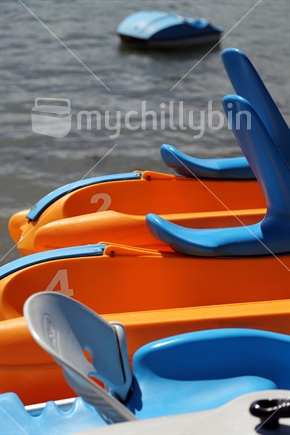 Close up detail of several colourful paddle-boats, all available for recreational hire, with a single paddle-boat anchored in the middle distance.