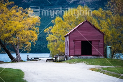 Old style shed in front of Trees in Autumn colours next to a lake.