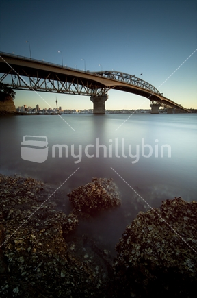 A long exposure looking back towards the city with the Harbour Bridge framing the city at dusk with the lights on the bridge illuminated, and rocks and oysters in the foreground. 