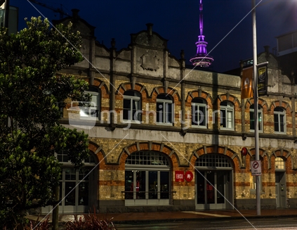 A brick arched windowed building with the Sky tower illuminated in the background