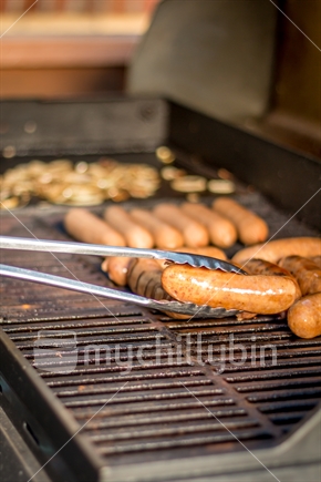 Sausages and onions being cooked on the BBQ, sausages being turned by tongs
