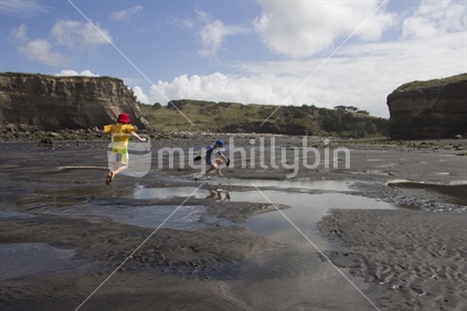 Two kids with sunhats jumping in puddles on secluded black sand beach