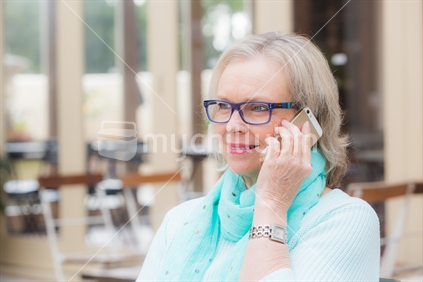 A blonde senior woman sits at outdoor cafe while listening to someone through her smart cell phone