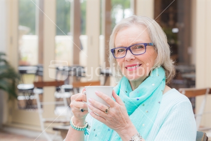 A beautiful middle aged blonde woman with glasses sits at a outdoor cafe with a drink