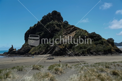 A view of lions rock on Piha  beach in Auckland.  With sand, sea, and the sky, and people on the sand enjoying a day at the beach. 